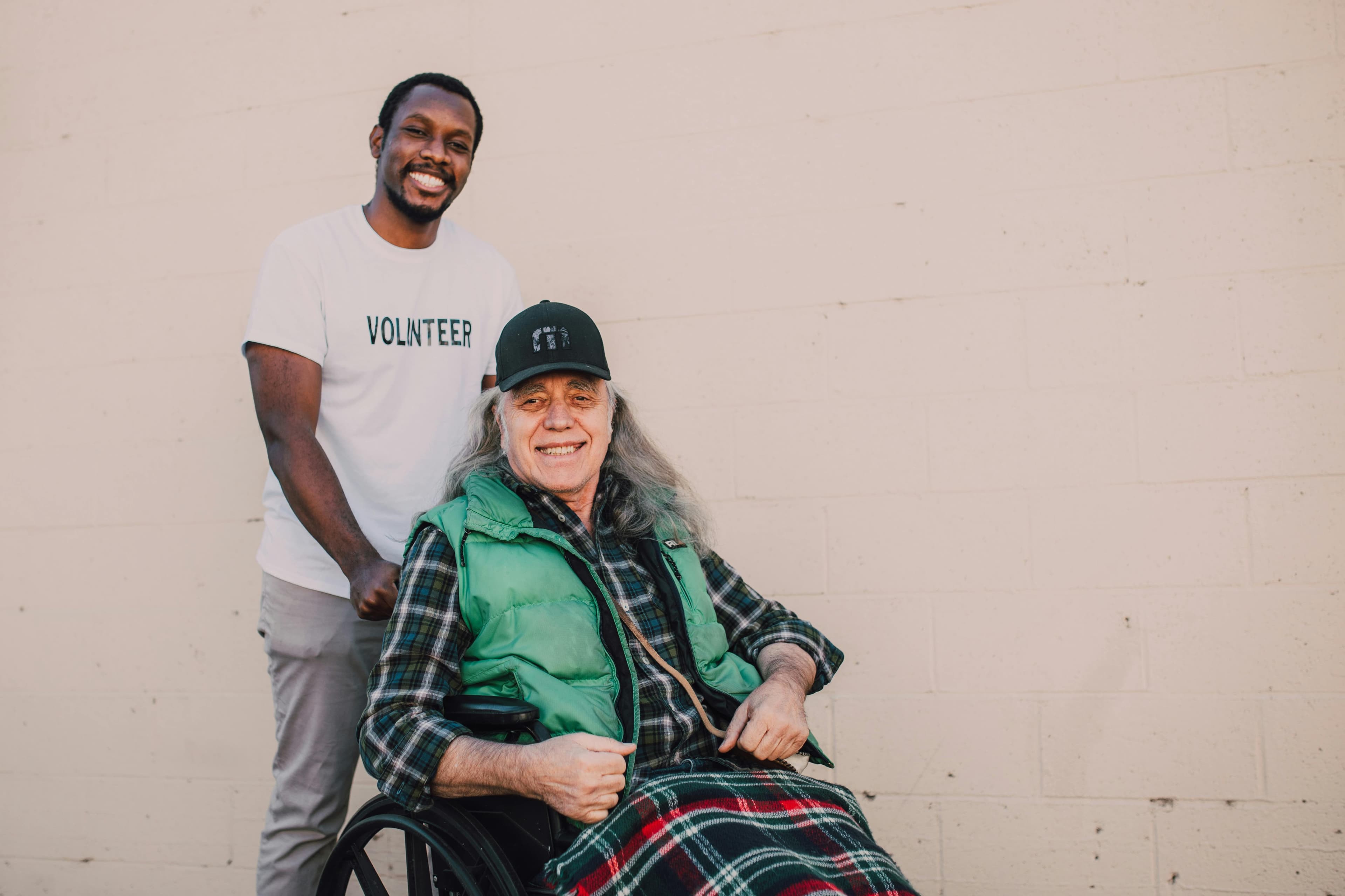 A volunteer helps a smiling man in a wheelchair outside.