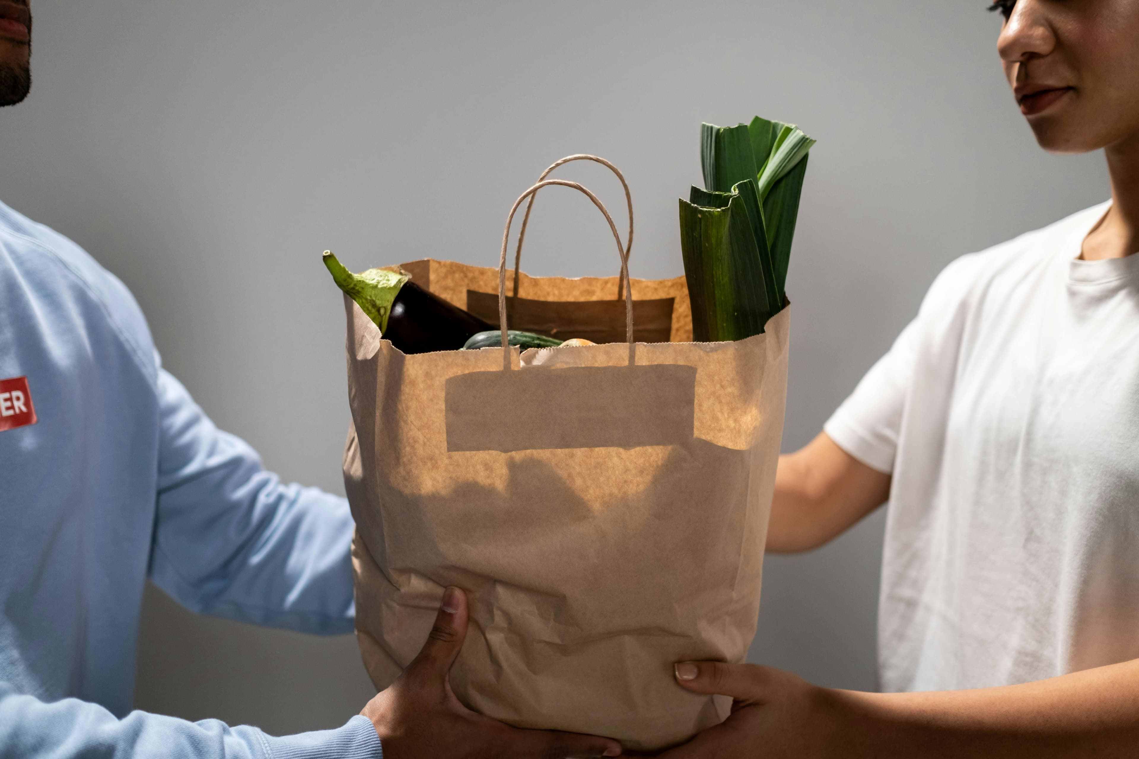 Two hands exchanging a brown paper grocery bag filled with fresh vegetables, including leeks and eggplant, in a neutral background.