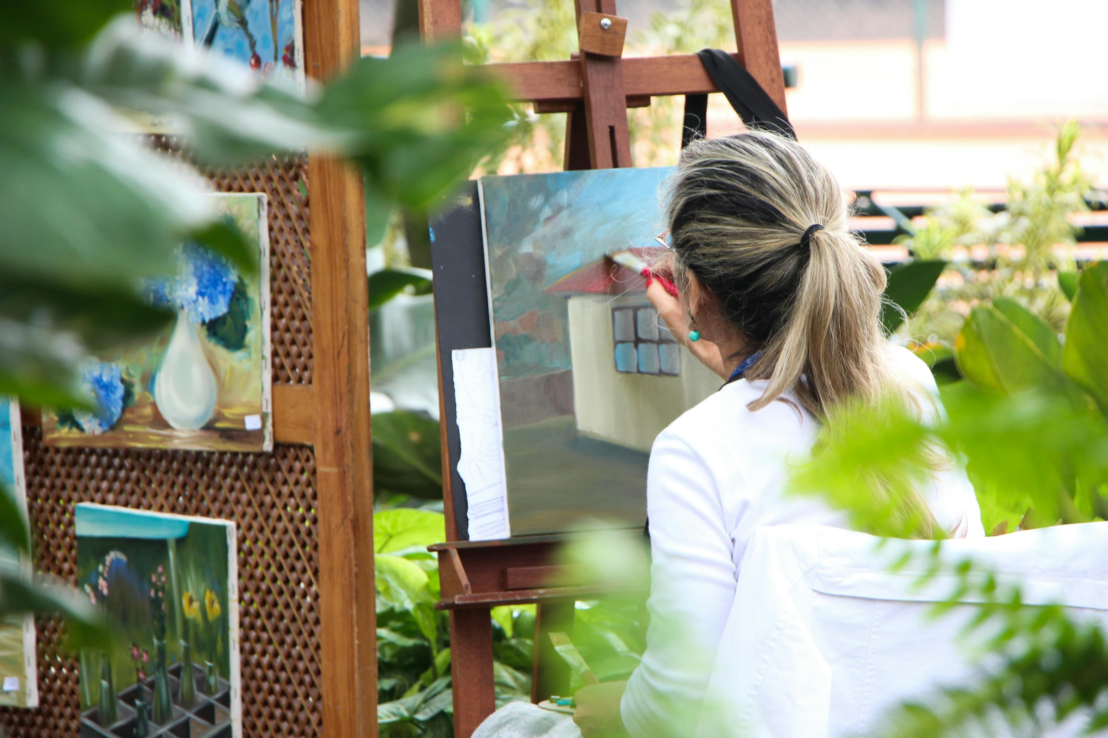 An artist with shoulder-length hair paints a house on canvas, surrounded by lush greenery and vibrant art on display.