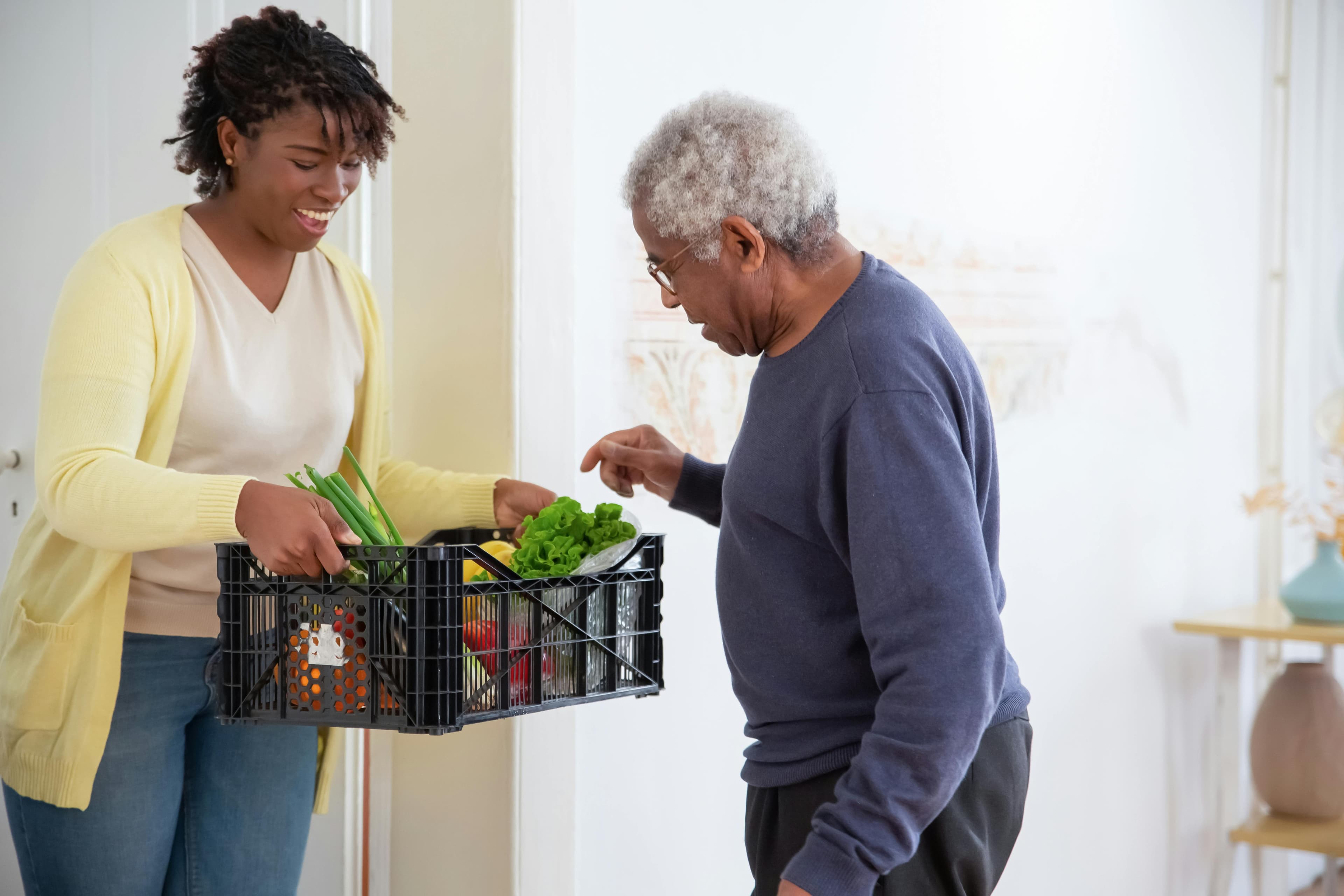 A woman in a yellow cardigan hands a crate of fresh vegetables to an older man, showcasing a moment of community support.