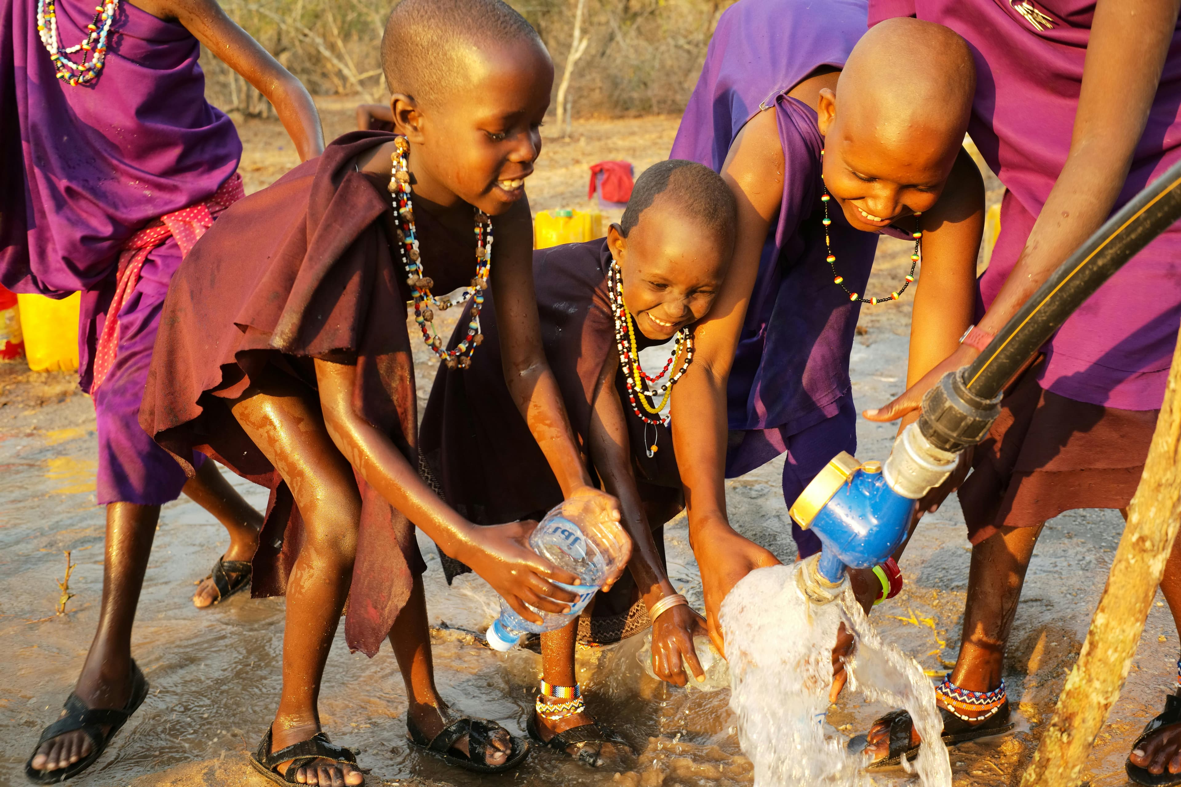 Children in traditional attire collect water from a pump, joyfully interacting around flowing water in a rural setting.