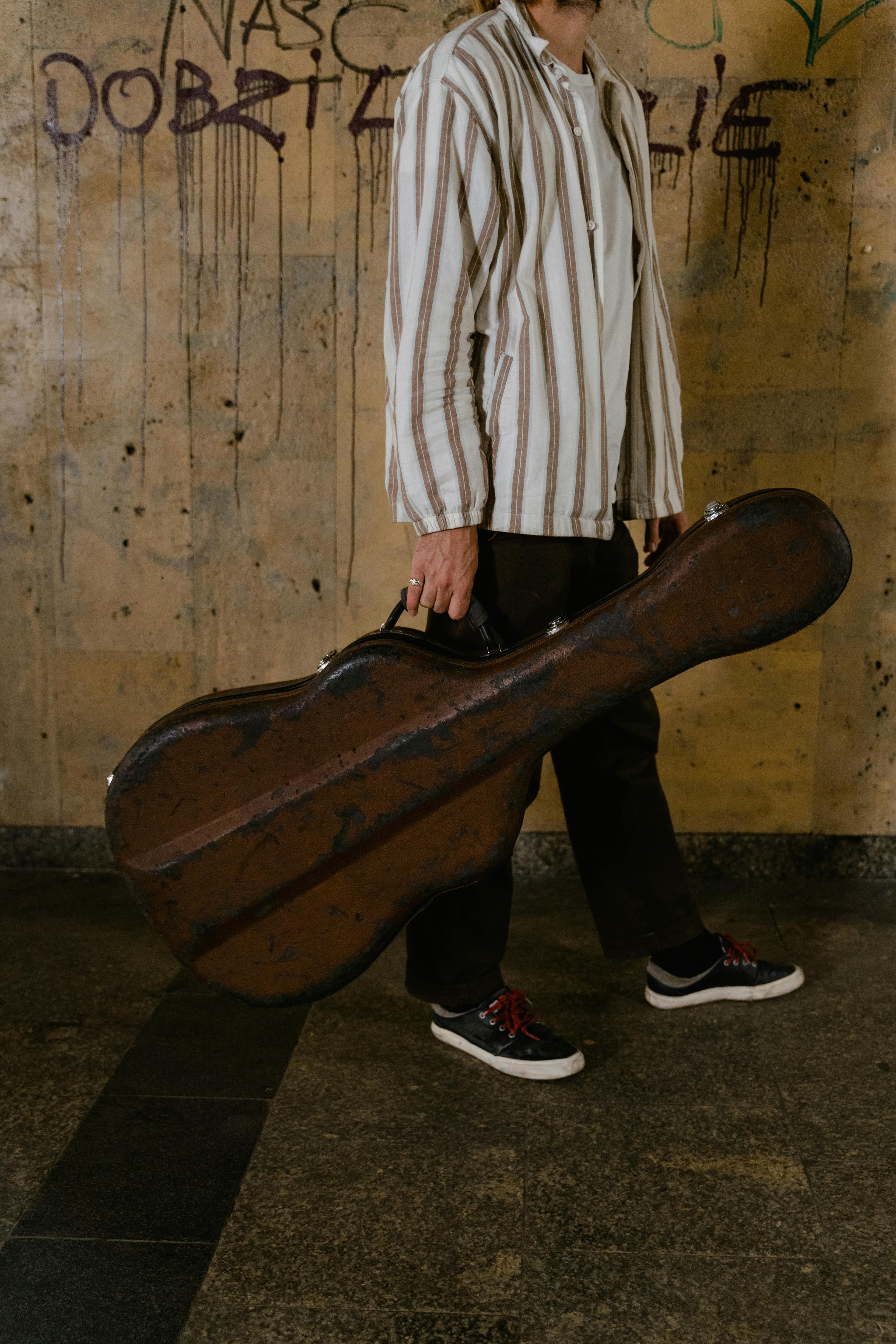 A person in a striped shirt holds a worn guitar case while walking on a gritty floor, with graffiti on the wall behind them.