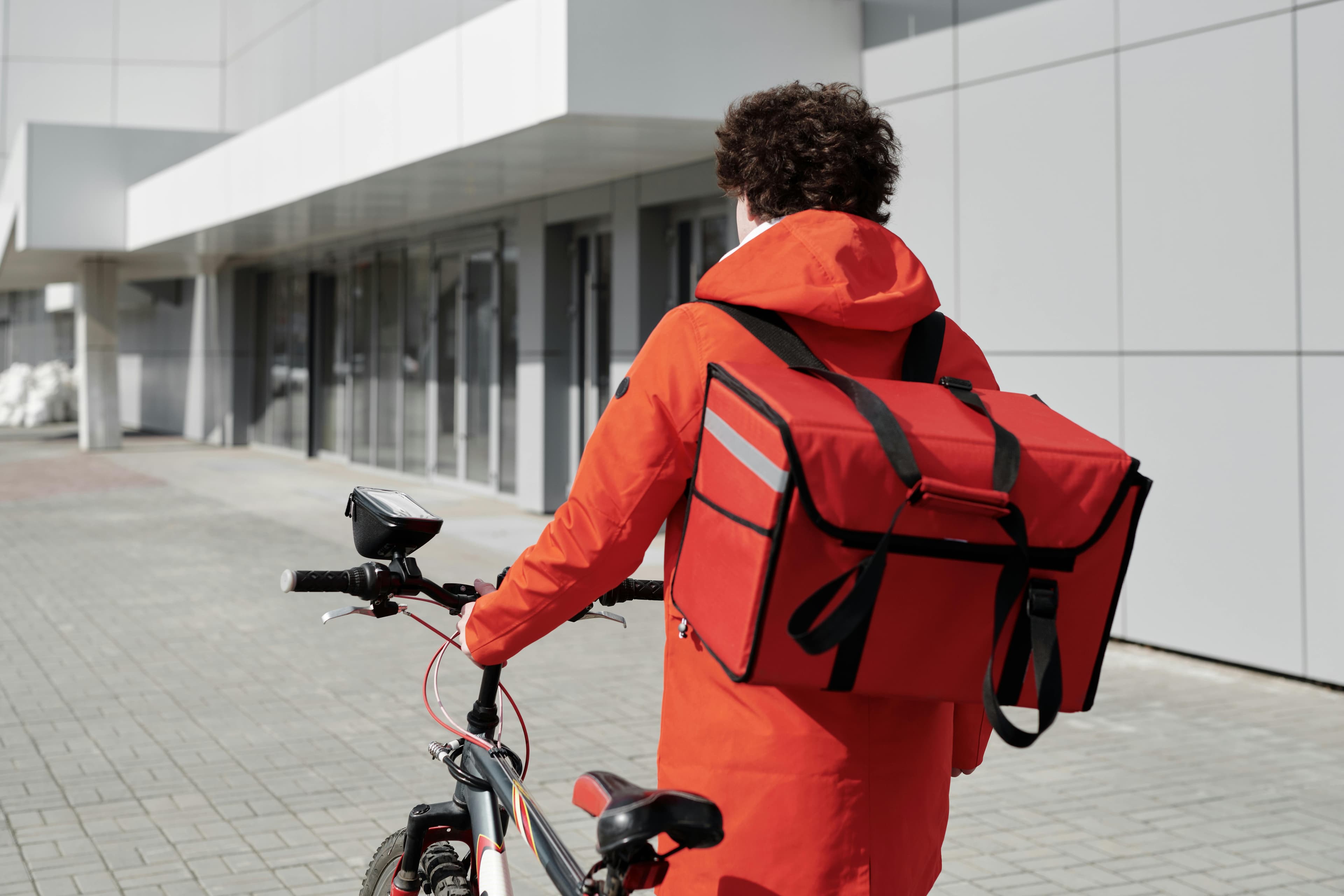 A cyclist in a bright orange jacket stands with a red delivery bag on their back, ready to ride in front of a modern building.