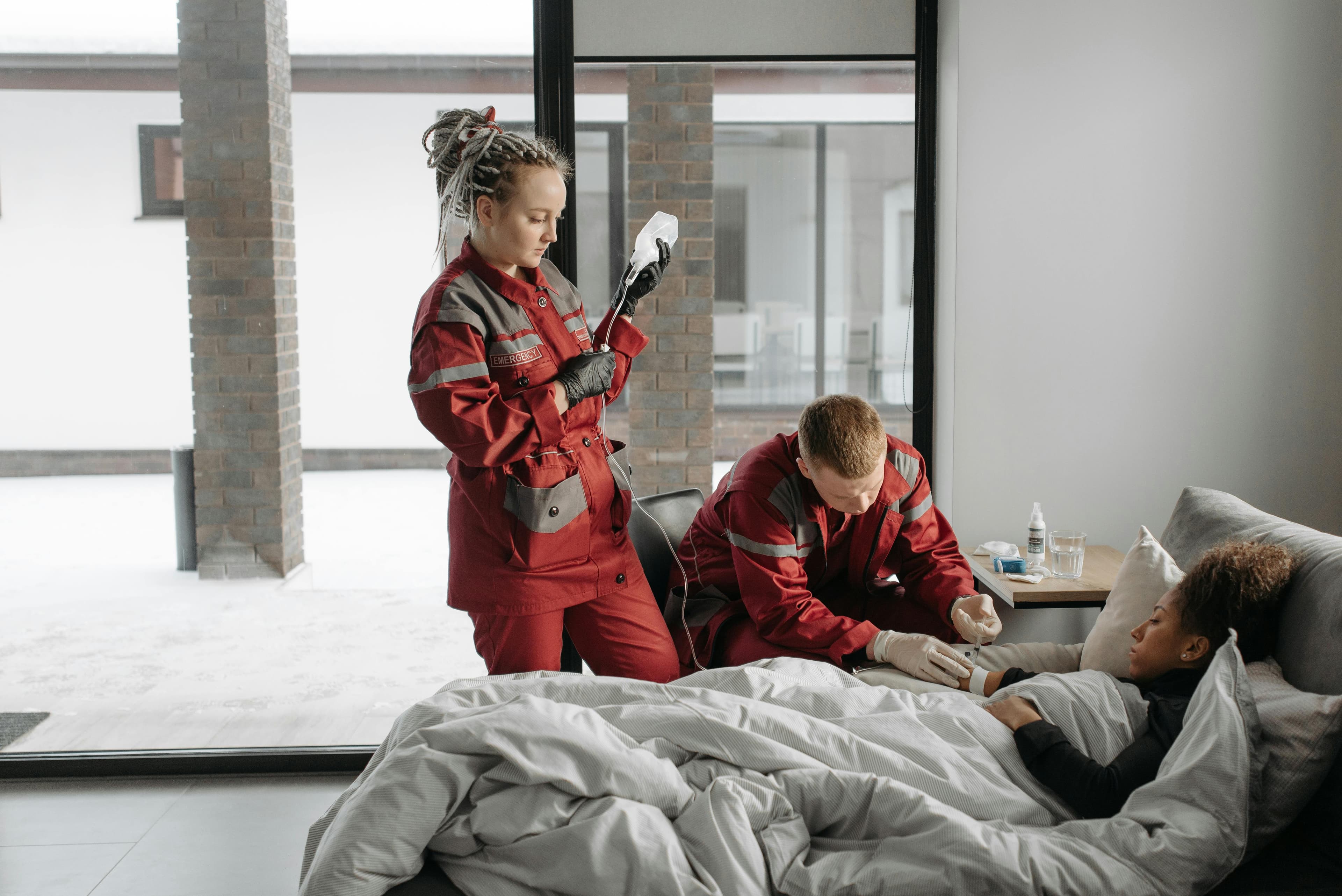 Two paramedics in red uniforms assist a patient lying under a gray blanket on a couch in a modern, well-lit room.