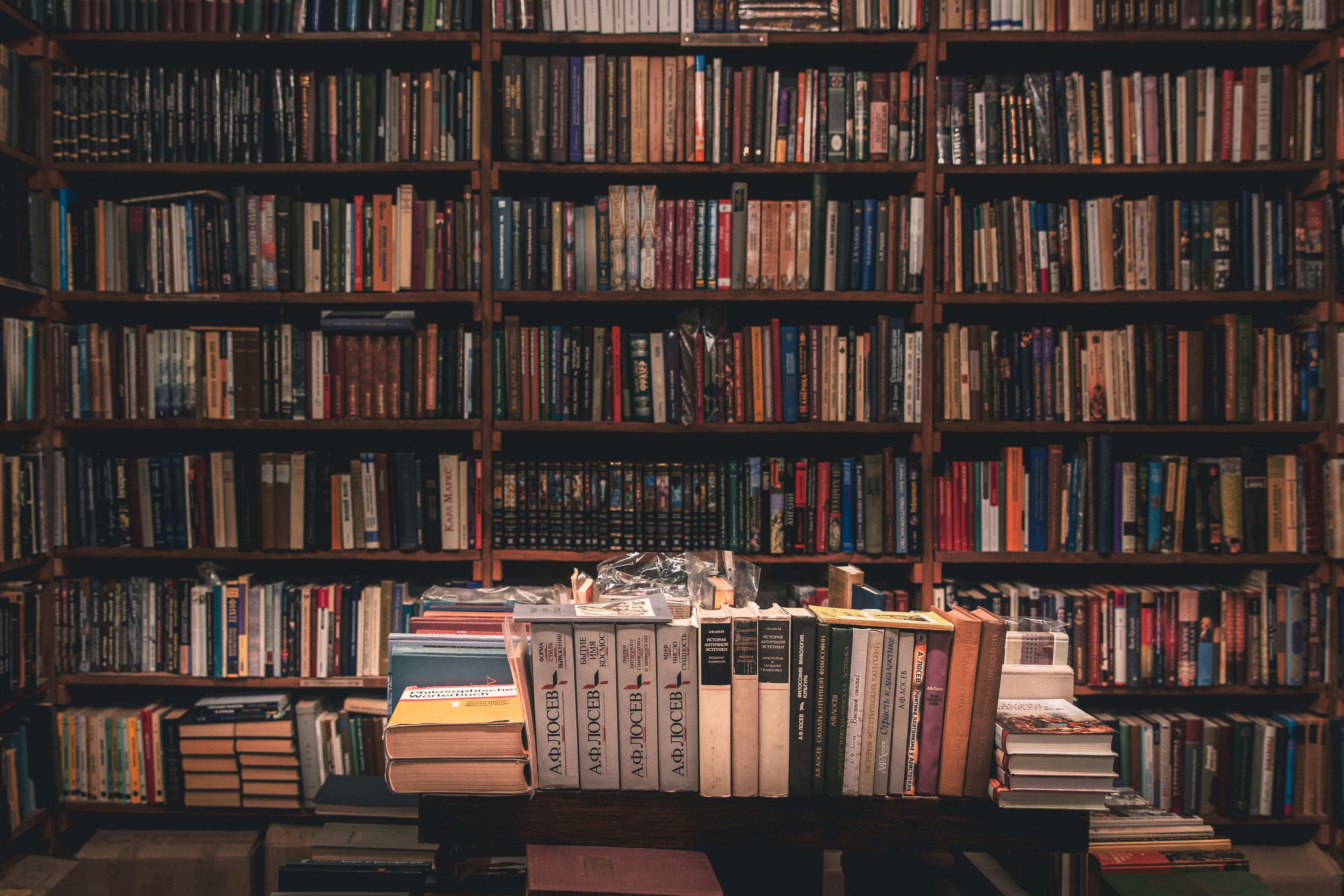 A cozy library with a wooden table stacked with assorted books, surrounded by shelves filled with various titles. Soft lighting enhances the atmosphere.
