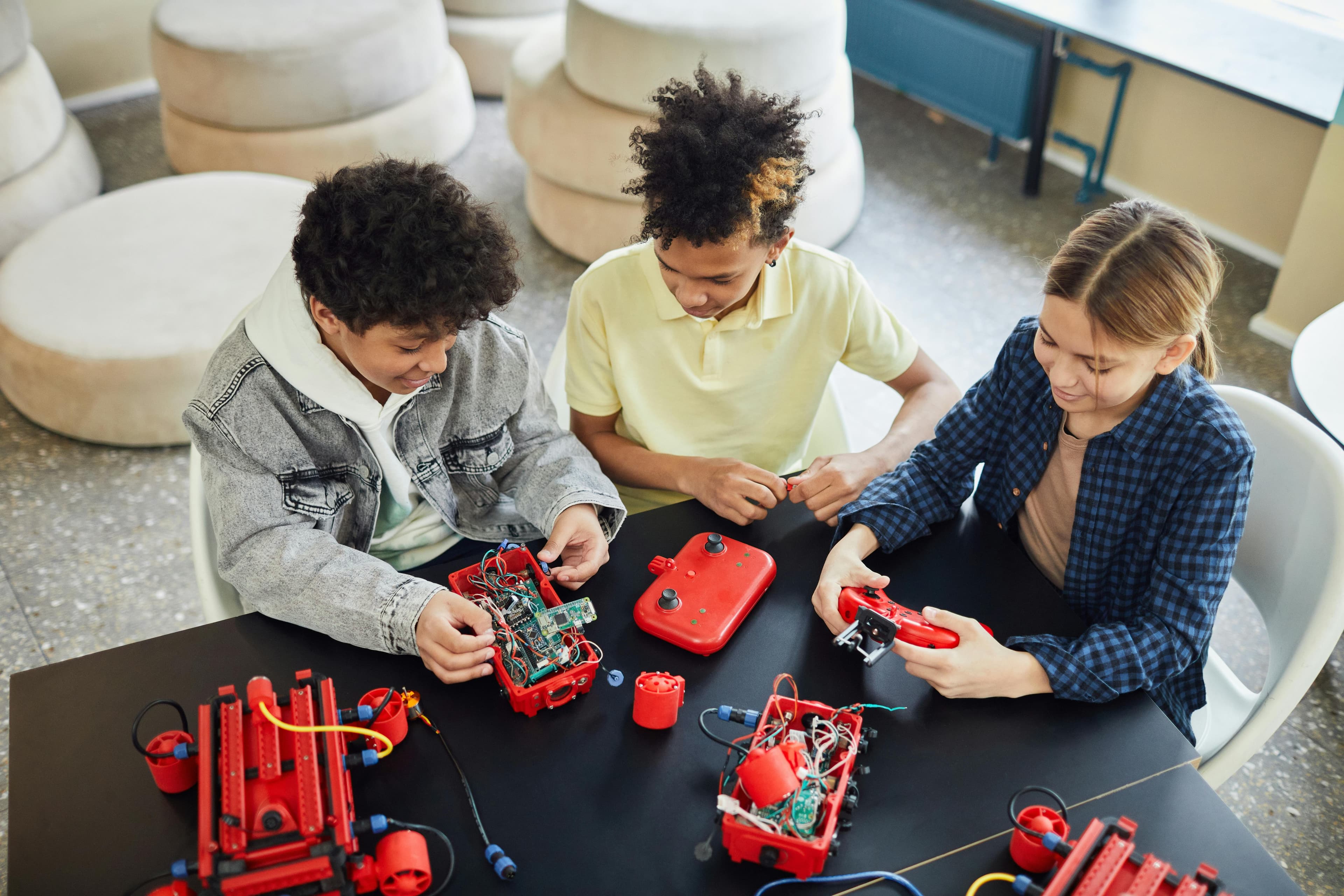 Three children collaboratively work on building and programming small robotic vehicles at a table filled with electronics and tools.