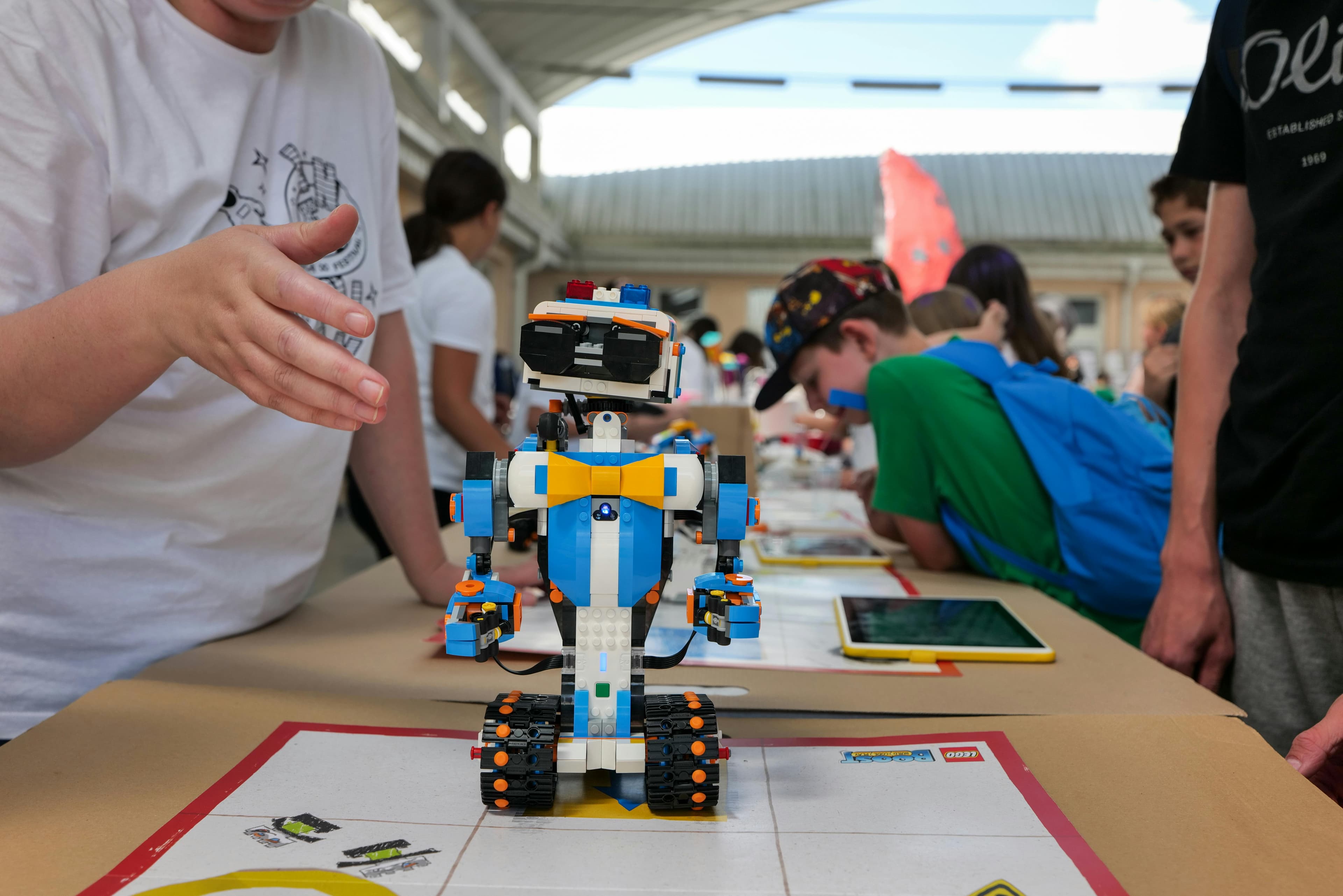 A colorful LEGO robot stands on a table, surrounded by children engaging in robotics activities, with tablets and drawings visible.